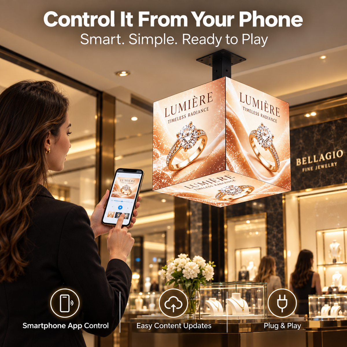 A woman in a jewelry store holds a smartphone app while controlling a hanging cube-shaped LED light display that shows a ring advertisement. Text at the top reads 'Control It From Your Phone' and the display reads 'LUMIÈRE ring light'.
